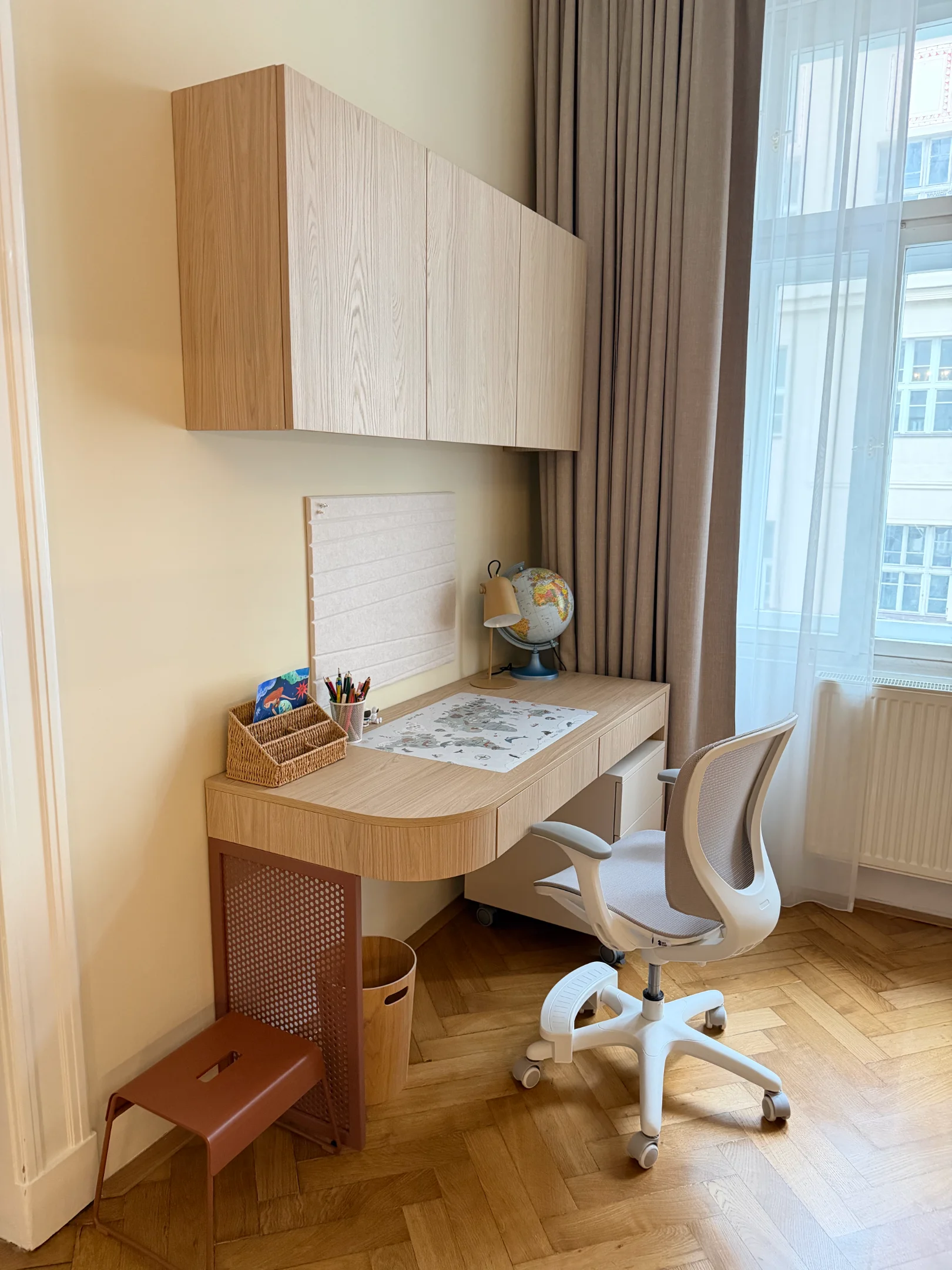 Světlý interiér s psacím stolem, skříňkou a židlí u okna.